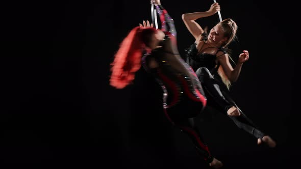 Two Gorgeous Slim Skilled Aerial Acrobats in Stage Costumes Spinning on Air Hoop at Black Background