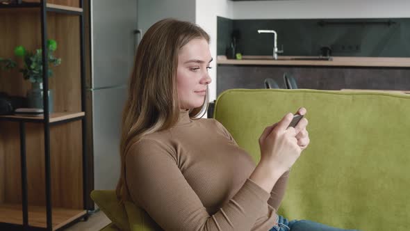 Pretty Young Woman Sitting on Couch at Home and Playing Game on Smartphone with Enjoy