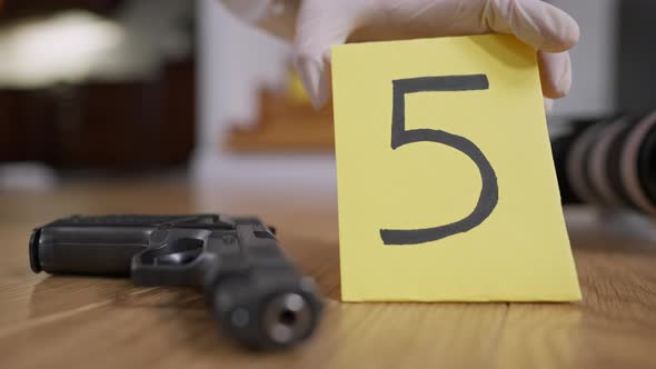 Closeup of Gun Lying on the Floor As Female Hand in Protective Suit Putting Numbered Marker at Crime