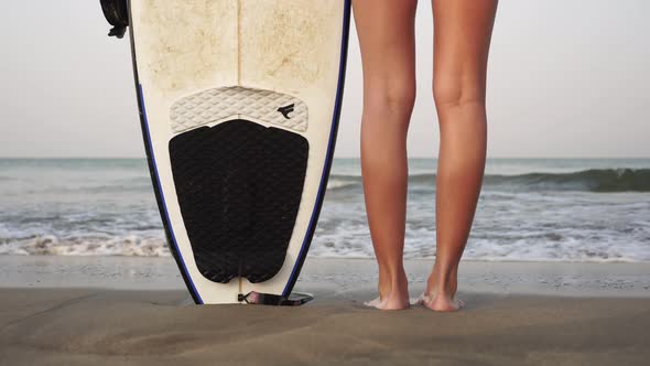 Young Woman Stands with a Surfboard on the Sandy Sea Coast