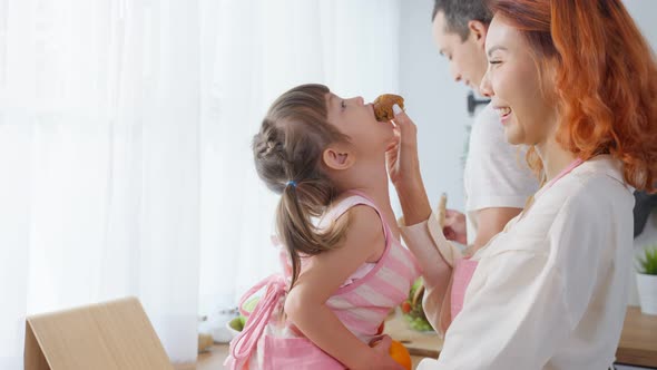 Asian attractive parents feed cookies to cute baby daughter in kitchen.