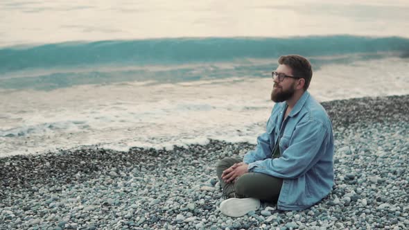 A Young and Bearded Man Sits on a Stony Beach and Enjoys the Sea View