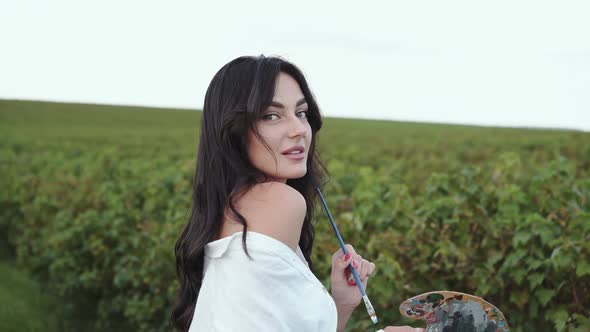 Brunette Smiles and Poses with Palette and Brush at Camera in Green Windy Field