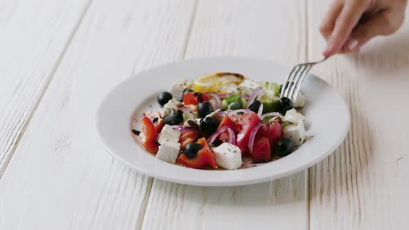 Greek Salad in a White Plate on a Wooden White Plate. Female Hand with Fork Close-up.