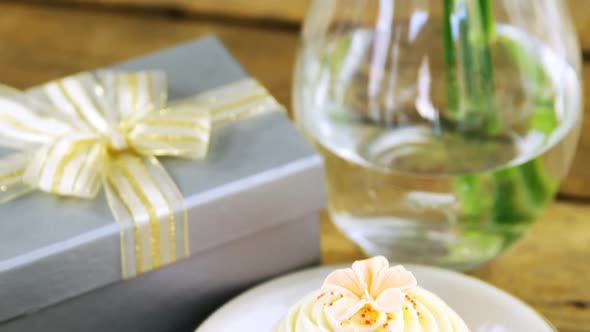 Gift box, flower vase and cupcake in plate on wooden plank