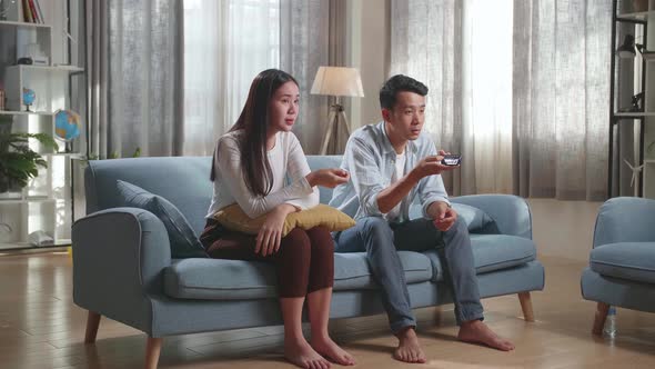 Young Asian Couple Is Watching Tv At Home. Man Switches Channels, Woman Selects Control