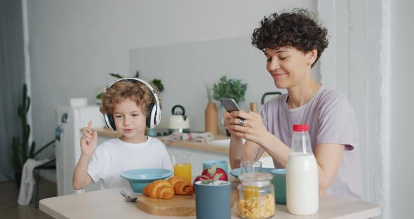 Young Lady Using Smartphone While Small Boy Enjoying Music at Kitchen Table