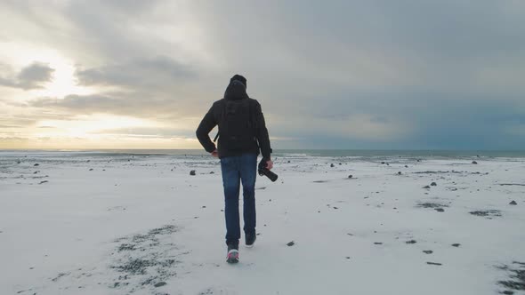 Young Man Traveler with Camera Runs to the Ocean on Snow Desert in Iceland