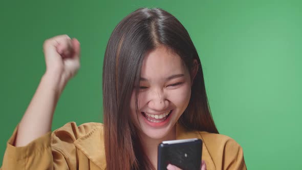 Young Asian Woman Celebrating With Mobile Phone While Standing On Green Screen In The Studio