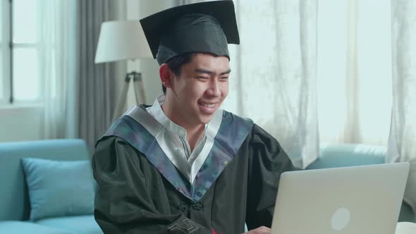 Asian Man Wearing A Graduation Gown And Cap Use Laptop Computer In Living Room
