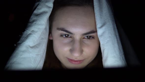 A Young Woman Lies Under a Blanket with a Tablet in Her Hands. A Woman in the Dark Uses a Pad