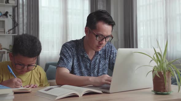 Asian Father Working With Laptop And Son Doing Homework At Living Room