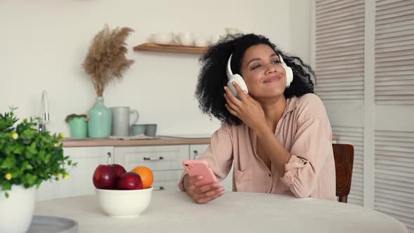 Cute African American Woman in Big White Headphones Listens to Music with Pleasure Using Smartphone