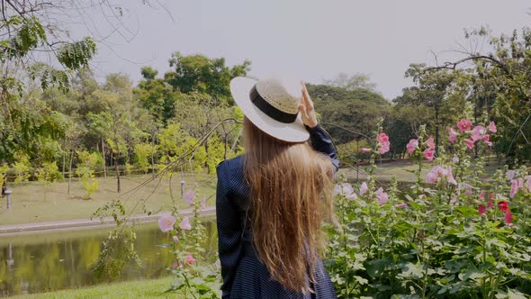 Travel Woman in Dress Walking in Garden with Flowers and Lake in Sunny Day