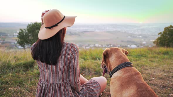 Back View of Adorable Girl with Hat Sits on the Meadow with Her Cute Dog