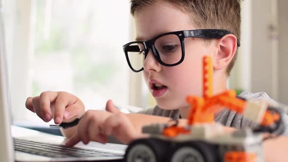 Smart boy in glasses programs a robot car and typing at a laptop at home.