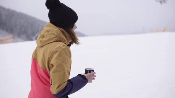 Girl with Takeaway Coffee Cup Over Ski Resort Background is Walking on the Snow