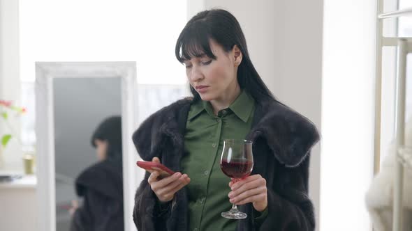 Portrait of Narcissistic Young Woman in Fur Coat Holding Glass with Wine Calling on Smartphone