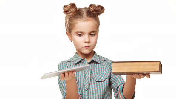 Small Serious Charming Caucasian Kid Girl in Plaid Shirt and Two Hair Buns Weighing Book and Tablet