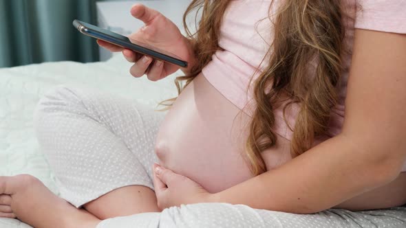 Closeup of Young Pregnant Woman Sitting on Bed and Browsing Internet on Smartphone