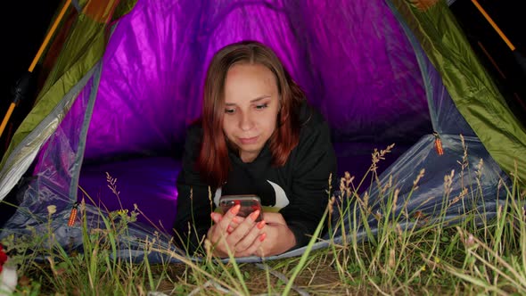 Young Woman Browsing Mobile Phone Lying in Tourist Tent at Night