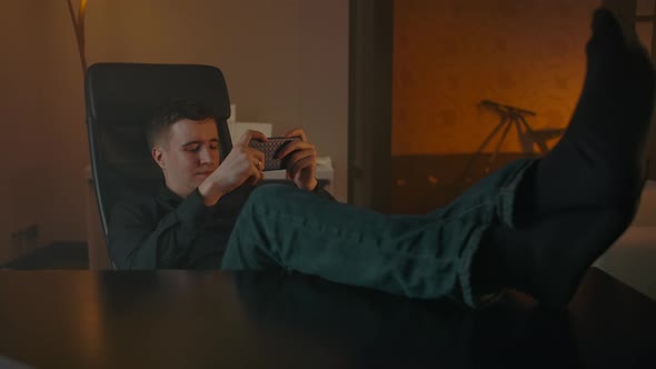 A Man Plays in the Phone Sitting at the Table Throwing His Feet on the Table