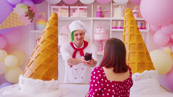Young man dressed as chef holding out muffin with lighting candle to woman in confectionery. 