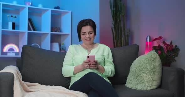 Senior Woman Sitting on Couch at Home Holding Invisible Smartphone in Hand and Chatting with Someone