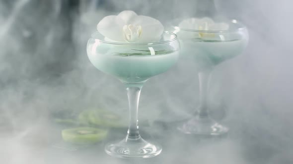 glass wine glasses with blue cocktail and white Orchid flower shrouded in smoke.