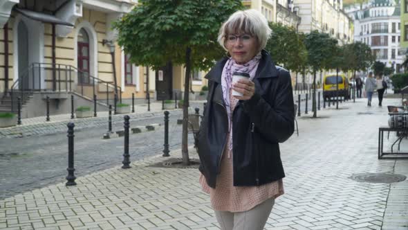Confident Caucasian Senior Woman in Eyeglasses Walking with Disposable Coffee Cup on City Street