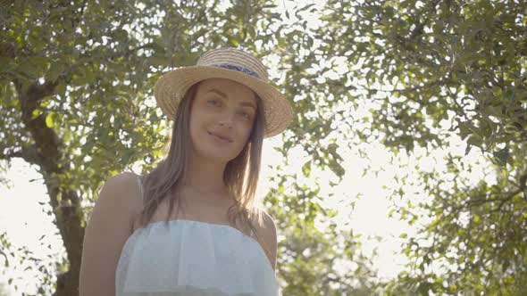 Attractive Positive Young Woman in Straw Hat and Long White Dress Looking at the Camera Smiling