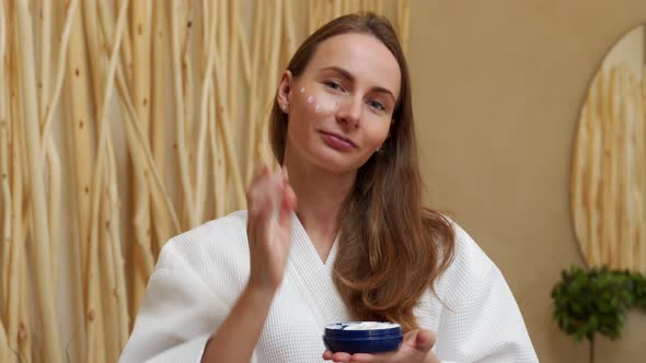 Smiling Beautiful Woman in a Bathrobe Puts a Skin Care Cream on Her Face