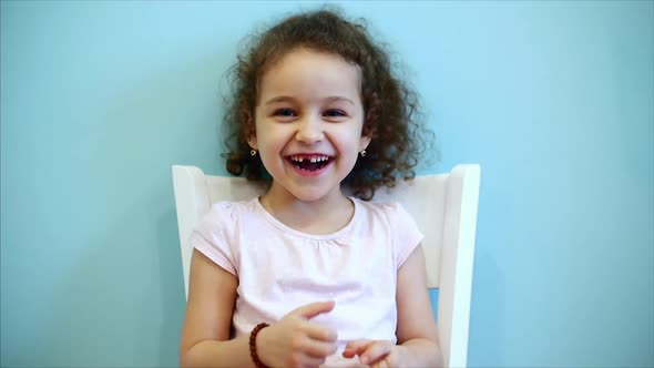 Cute Little Beautiful Funny Baby with Red Curly Hair Girl Sitting on a White Chair on a Blue