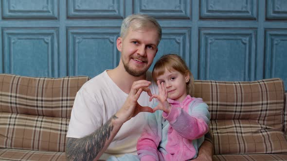 Father and Child Daughter Kid in Pajamas Sit on Couch Smiling Making Sign of Shape Heart Near Chest