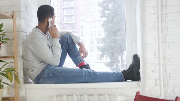 AfroAmerican Man Sitting at Window and Talking on Mobile Phone