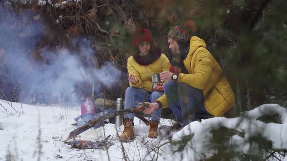 Young Man and Woman Drink Tea By the Fire in the Winter Forest