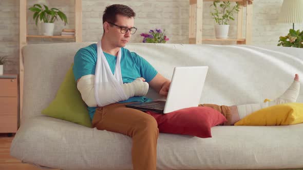 Portrait of a Young Man with a Broken Arm and Leg Sitting on the Couch and Uses a Laptop