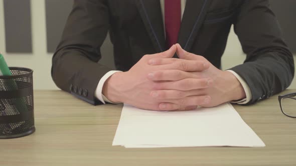 Unrecognizable Man in the Suit Sitting at the Table with Crossed Fingers in Front of Blank Papers