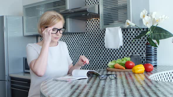 Woman Is Sitting in the Kitchen at the Table and Reading a Recipe for a Dish