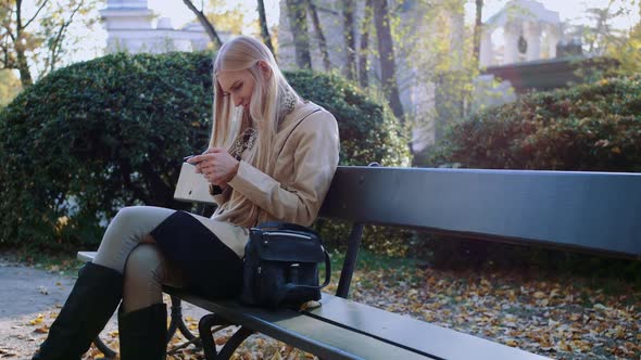 Girl Communicates in a Smartphone in a City Park on a Bench.