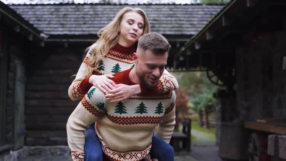 Happy young lovely couple having fun near wooden house in the park. Man piggybacking girl, running h