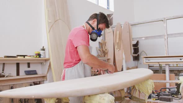 Caucasian male surfboard maker wearing a face mask and preparing to polishing a surfboard