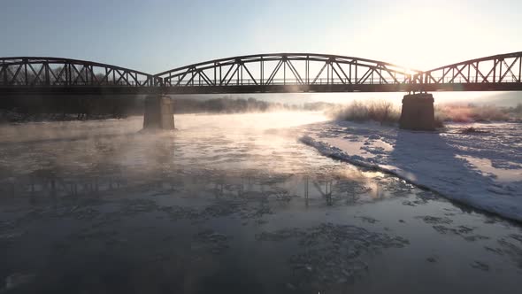 Cinematic Drone Shot Flies Forward Over the Frozen River and Under the Red Steel Bridge at Sunrise