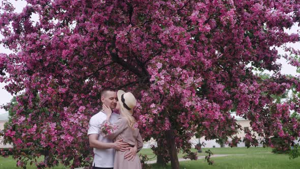 Romantic Beautiful Woman in a Hat Stands with a Man at a Cherry Blossom Tree in the Summer in the