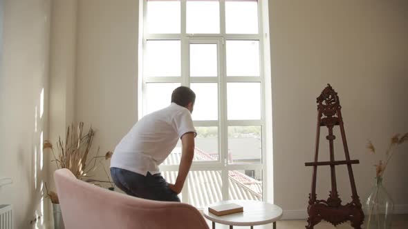 A man in spacious room gets up from the sofa and goes to think to the big window