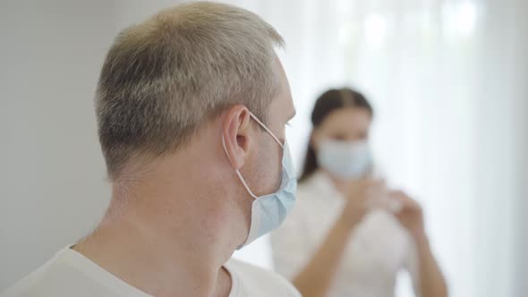 Close-up of Scared Male Patient in Face Mask Looking Back at Nurse Filling Syringe and Turning To