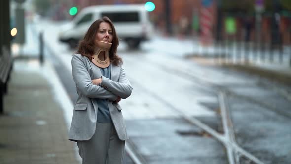 a Middleaged Woman with an Orthopedic Collar Around Neck Has Arms Folded and Stands Against the