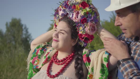 Adult Gardener in a Helmet and Overalls Walks Up To a Cute Plump Girl with a Wreath on Her Head