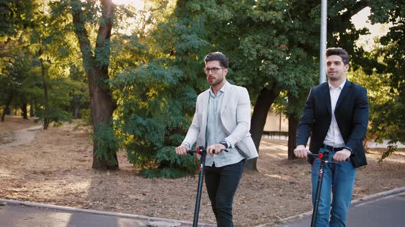 Two Handsome Young Businessman Riding an Electric Scooters on the Road in City Park Slow Motion