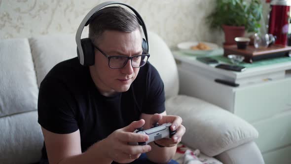 Young Man in Glasses and Headphones Plays Console Intently and Sits on the Couch at Home